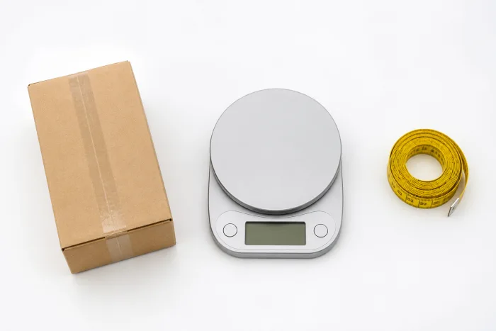 Balança ao lado de uma caixa de papelão com fita métrica, representando o cálculo de frete pelos Correios
